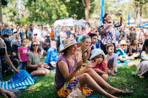 A crowd at an outdoor community event sitting on grass applauding