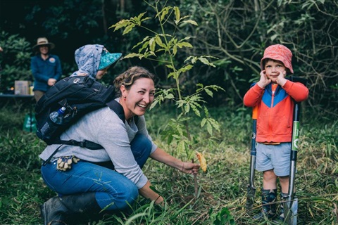 The City of Coffs Harbour's Reslience and Recovery service is portrayed with a picture of a woman planting a tree and smiling at the camera. She has a baby in a carry pack on her back and a young child helping her.